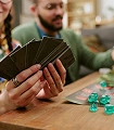 Op de afbeelding is een groep mensen te zien die deelneemt aan een rollenspel of kaartspel. Ze zitten aan een houten tafel in een gezellige, goed verlichte kamer met boekenplanken en huiselijke elementen op de achtergrond. De sfeer is vriendelijk en ontspannen, wat een gevoel van luchtig vermaak en gezelligheid oproept. Het kleurenpalet van de afbeelding is warm en rijk, met zachte tinten bruin, groen en rood.